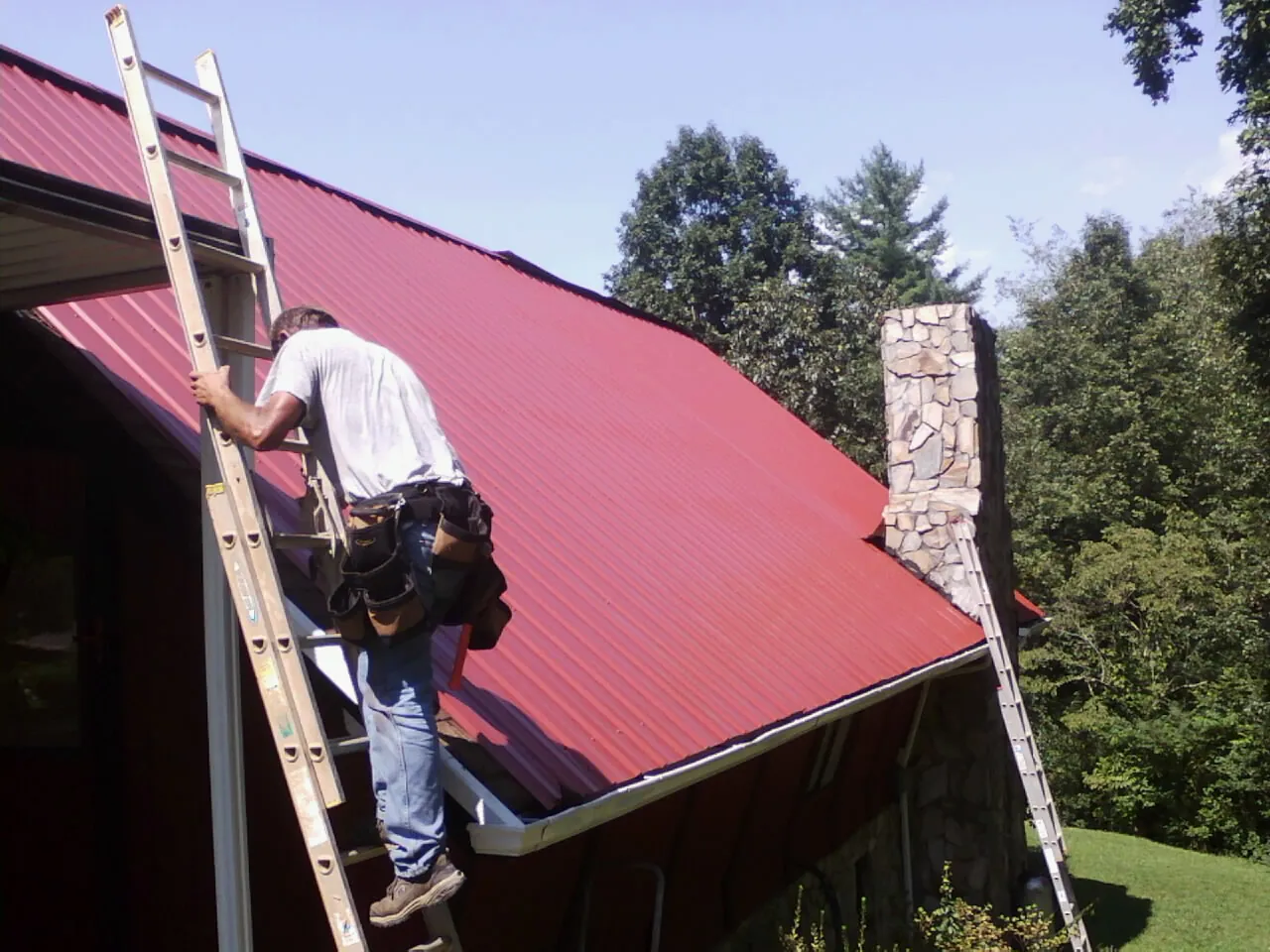 Roofer on a ladder beside a red metal roof and stone chimney — Soffit & Fascia Repair by Roofing Brothers in Coralville